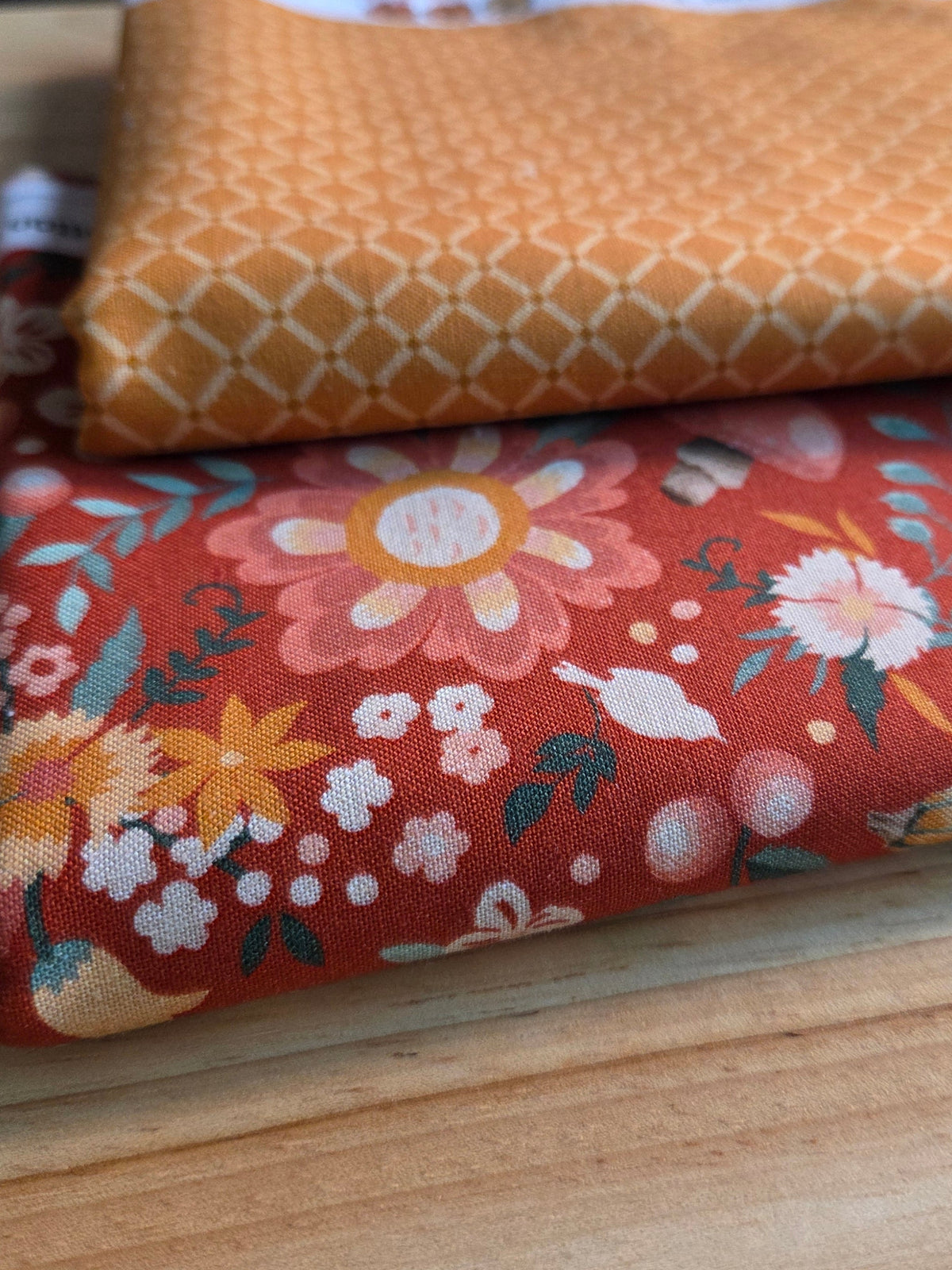 Two folded fabrics stacked on a light wood surface. The top fabric is a warm orange with a cream geometric lattice pattern and a white lace trim.