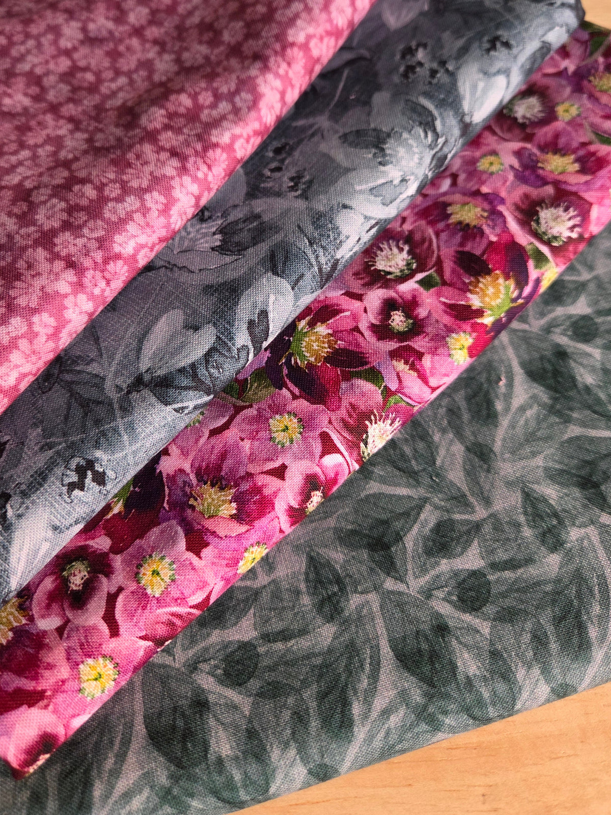 Four folded fabrics stacked diagonally on a wooden surface, featuring pink floral and gray botanical patterns.