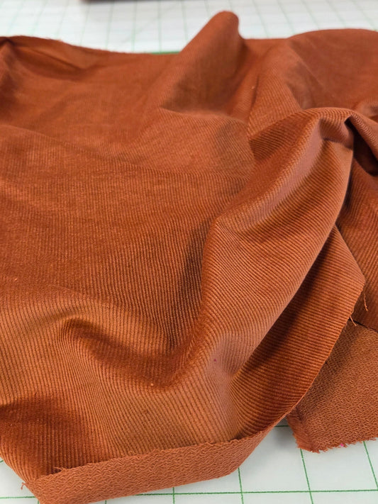 A close-up of rust-orange corduroy fabric draped on a white cutting mat, with a matching rolled piece of fabric visible in the background. baby wale deadstock