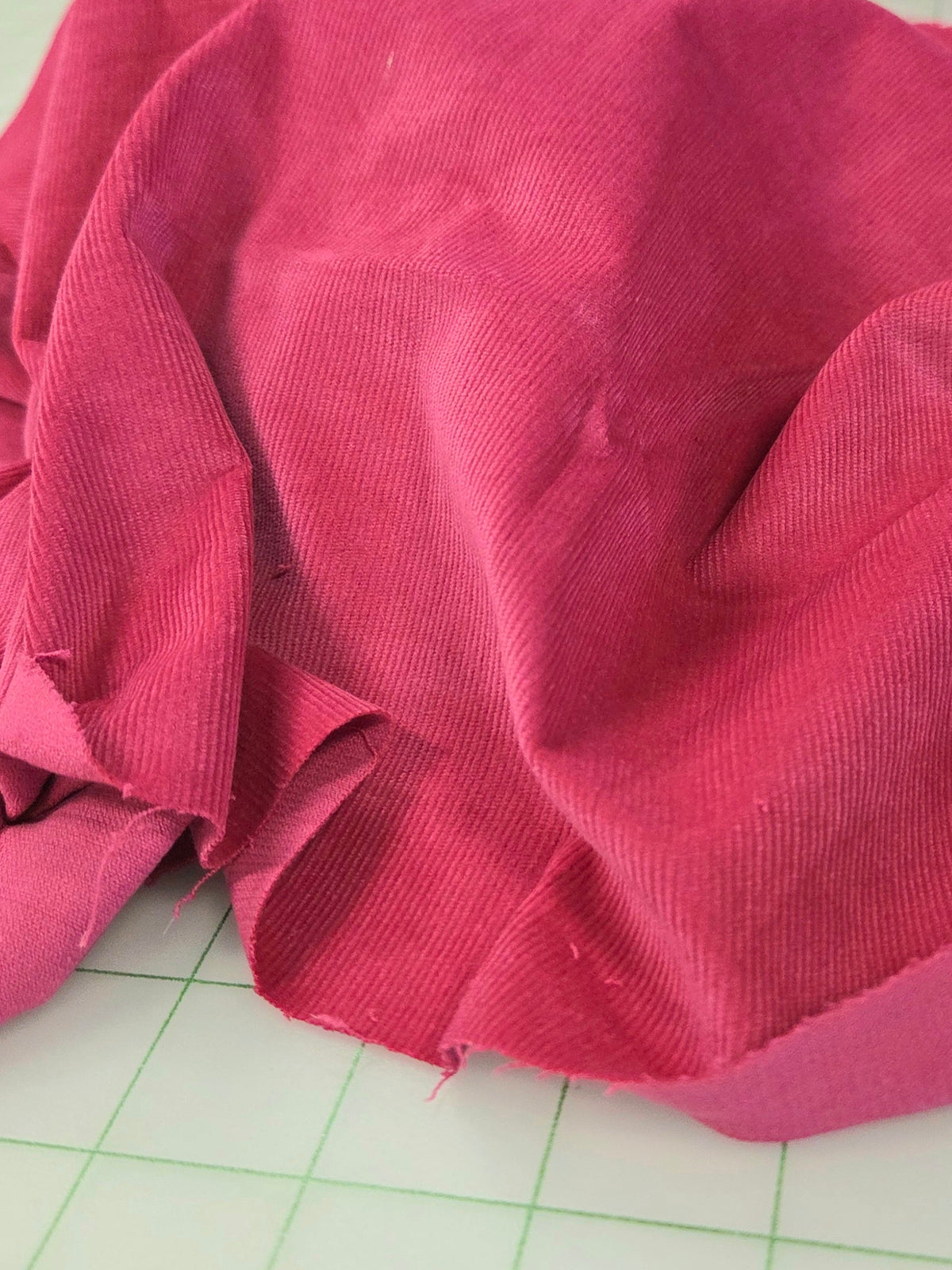 A close-up of bright pink fabric draped on a green-grid cutting mat, with a roll of orange-brown fabric in the background. corduroy deadstock by the yard baby wale magenta. LA Finch