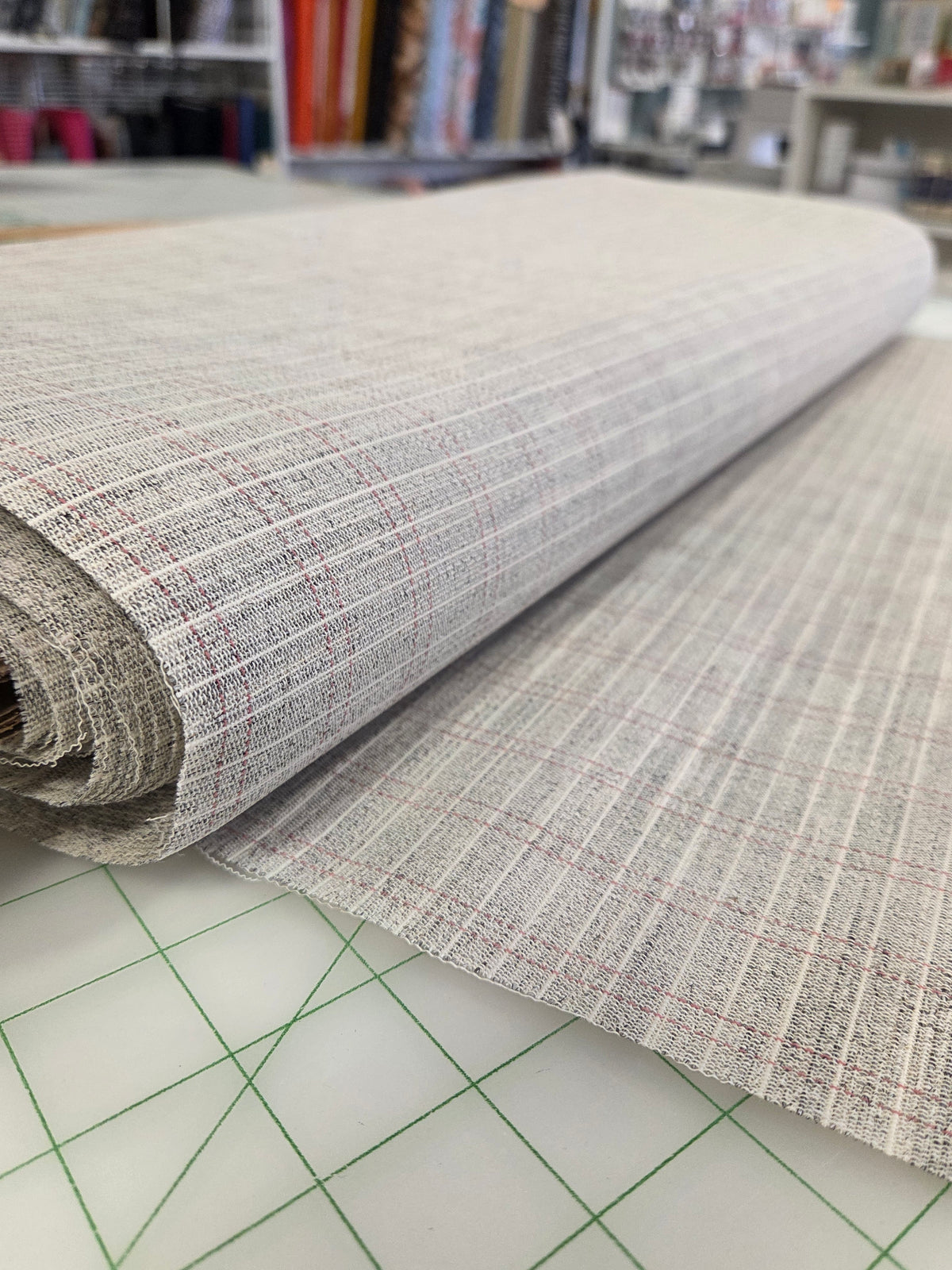 A close-up of a rolled piece of woven fabric with a light gray and white textured pattern and subtle red lines, laid over a green-grid cutting mat in a craft room setting. sewin horsehair canvas tailoring by the yard