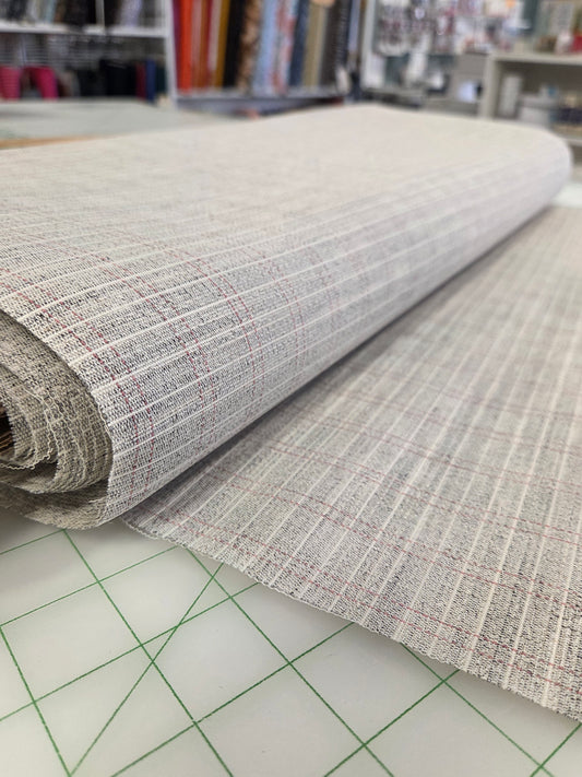A close-up of a rolled piece of woven fabric with a light gray and white textured pattern and subtle red lines, laid over a green-grid cutting mat in a craft room setting. sewin horsehair canvas tailoring by the yard