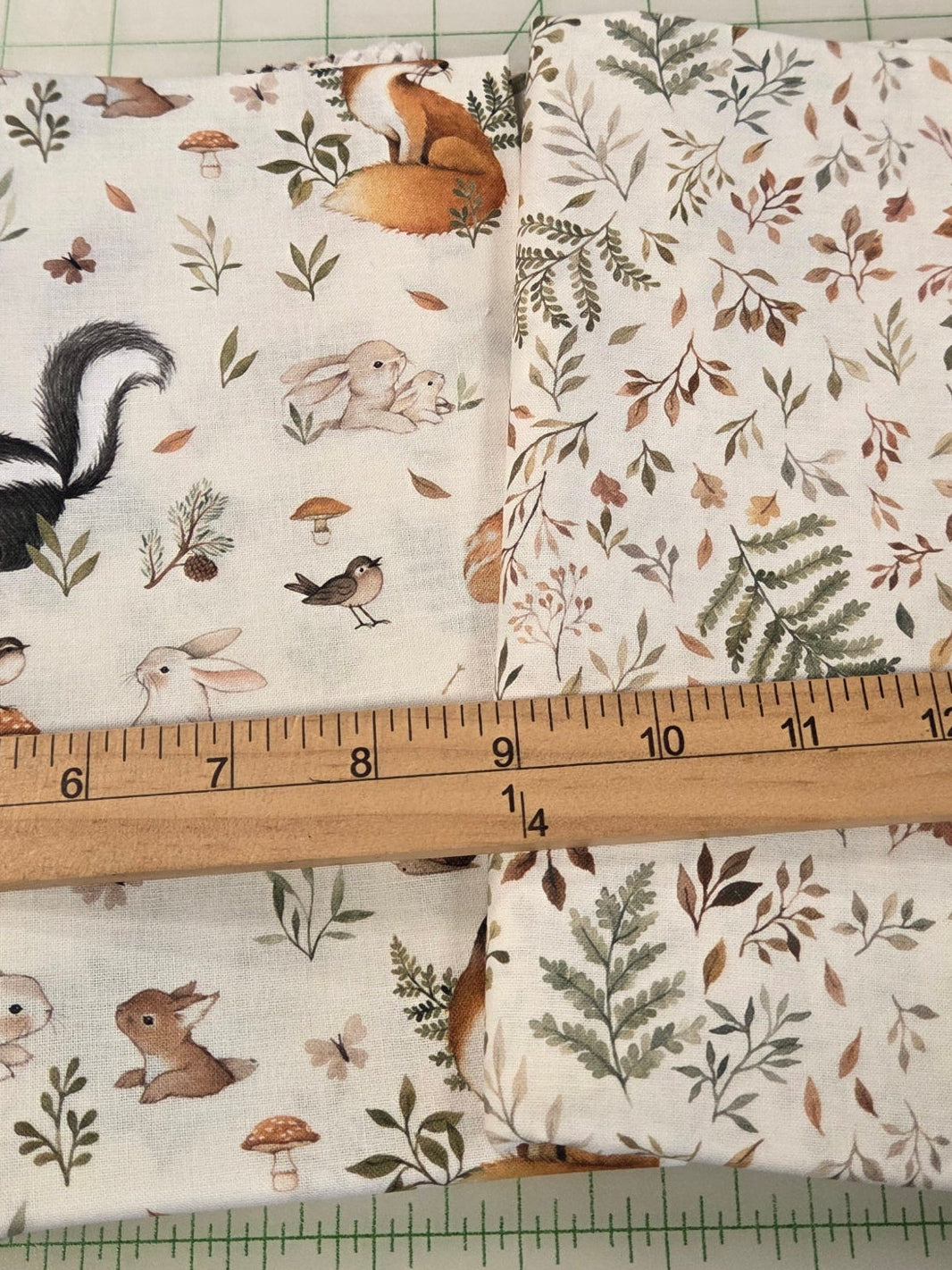A folded piece of fabric with a woodland animal and floral print pattern, laid on a white cutting mat with a wooden ruler across it for scale.
