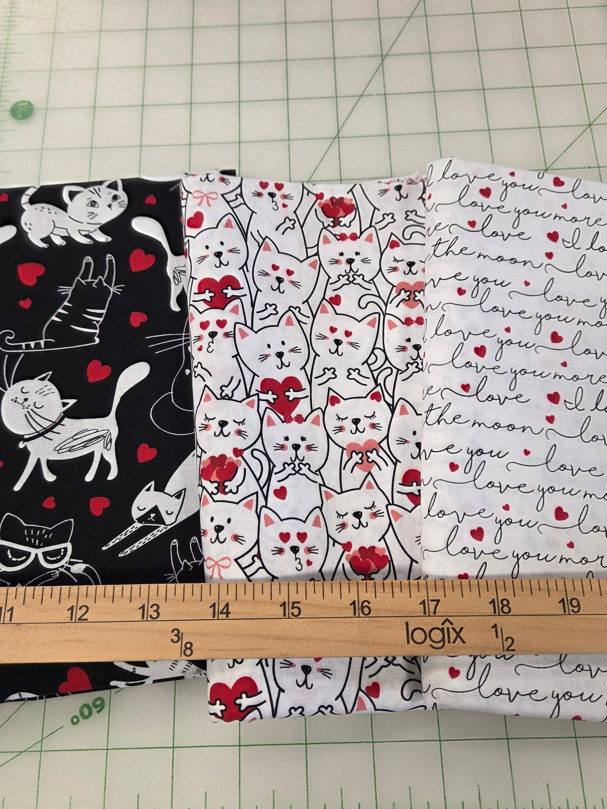 Three patterns of fabric swatches featuring cats and 'I love you' text, displayed on a green cutting mat with a wooden yardstick for scale.