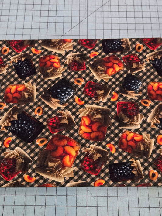 A rectangular fabric or paper sheet with a repeating fruit-and-basket pattern on a dark checkered background, photographed on a gray gridded cutting mat. The pattern includes oranges, reds, blackberries, and blueberries in baskets and jars.