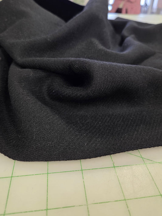 A close-up of draped black knit fabric resting on a green-lined cutting mat, with a softly blurred indoor workspace in the background.
