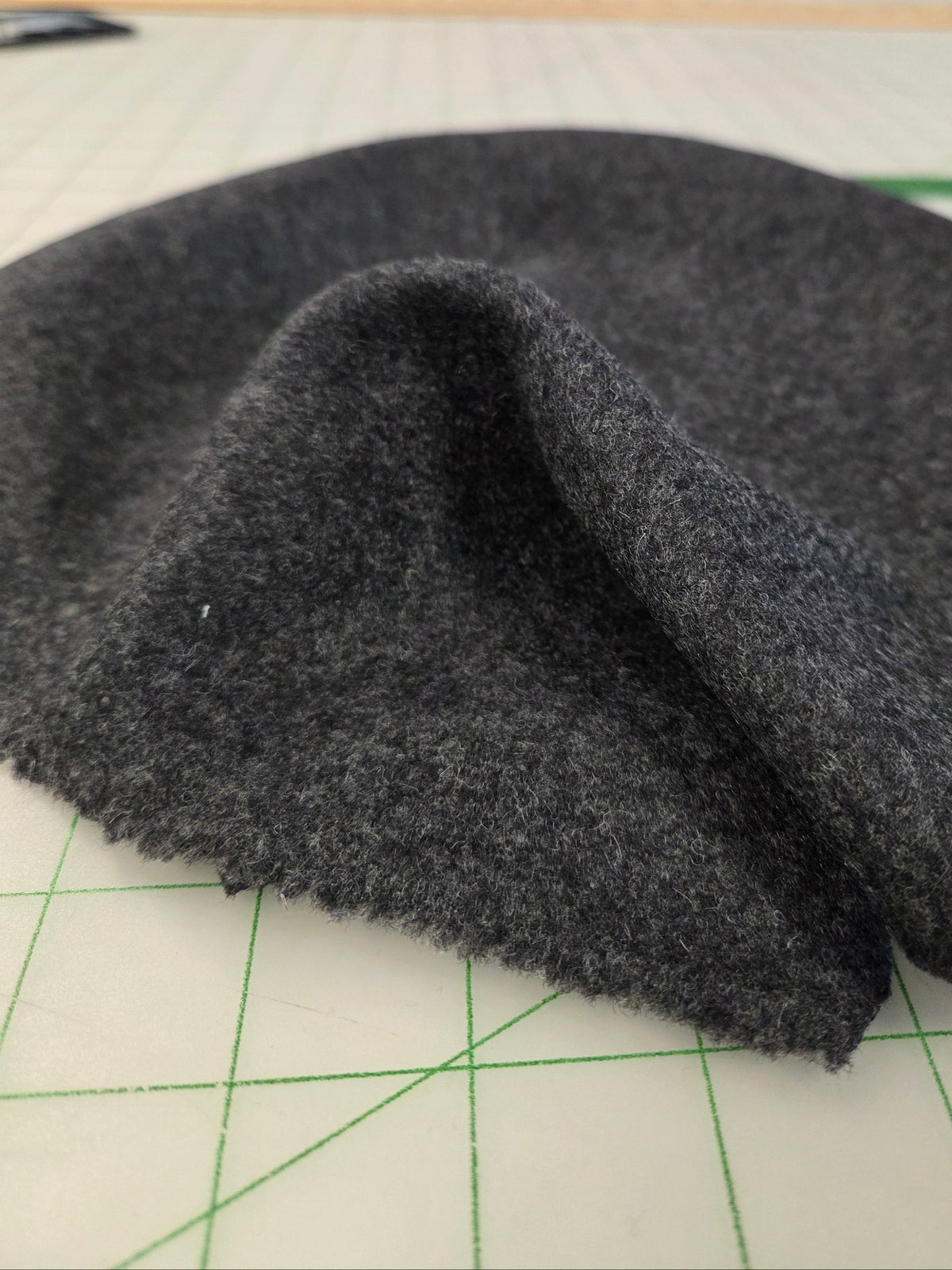 A close-up of a dark gray, textured fabric or felt material draped over a green-lined cutting mat, with a blurred background showing shelves and items.
