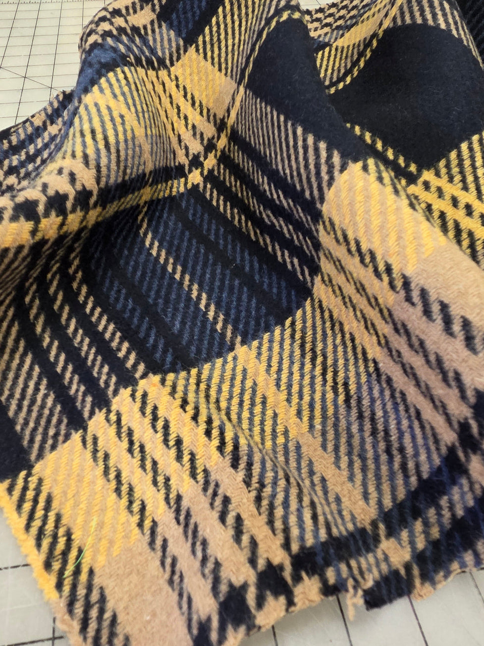 A close-up of a plaid woven textile with navy, beige, and yellow stripes, laid on a white gridded cutting mat. Wool Coating Italian