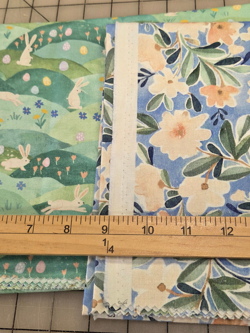 Two folded fabric bundles with floral and bunny prints laid on a gridded cutting mat, with a wooden ruler standing vertically to show measurements.