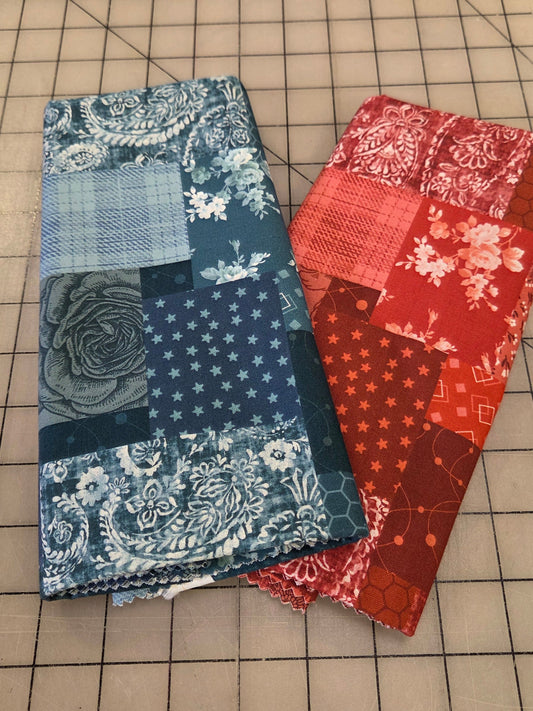 Two handmade patchwork fabric pouches or wallets stacked on a beige cutting mat with grid lines. The top pouch is made of red and orange floral and patterned fabrics, and the bottom pouch is made of blue and navy floral, paisley, and polka dot fabrics.