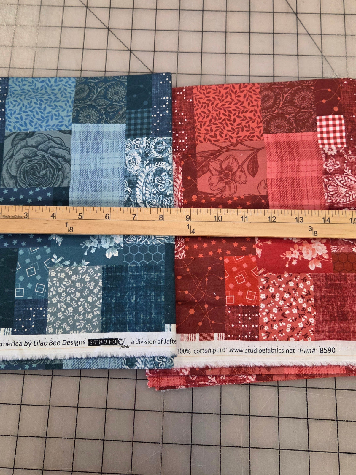 Two folded fabric bundles with patchwork patterns in red and blue, laid flat on a gridded cutting mat with a wooden ruler placed vertically between them.