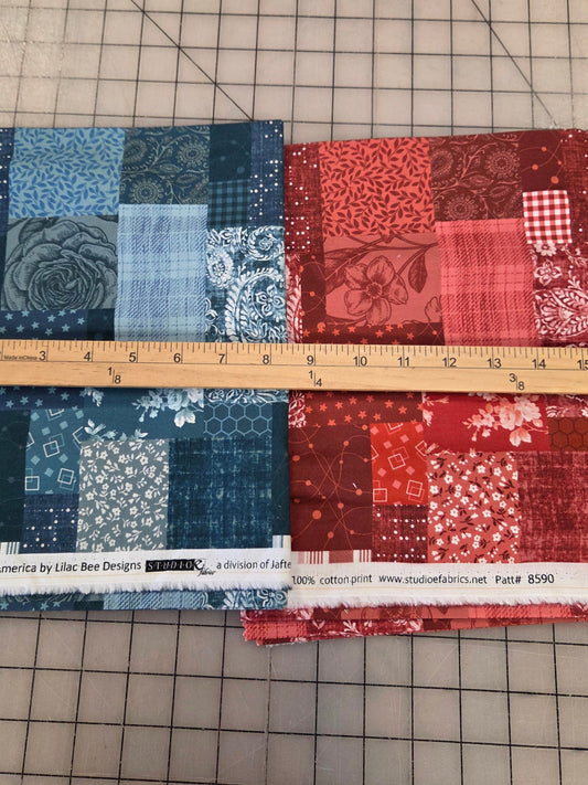 Two folded fabric bundles with patchwork patterns in red and blue, laid flat on a gridded cutting mat with a wooden ruler placed vertically between them.