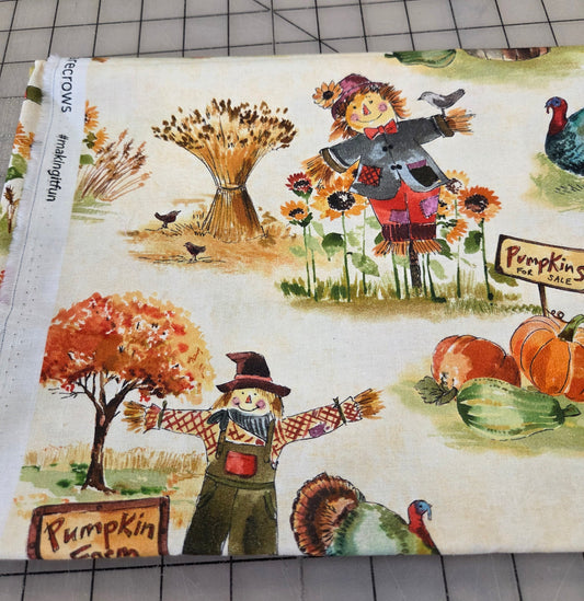 A close-up of a fabric or paper print featuring a fall harvest scene with scarecrows, pumpkins, sunflowers, a turkey, and autumn trees on a cream background, photographed on a gridded cutting mat.