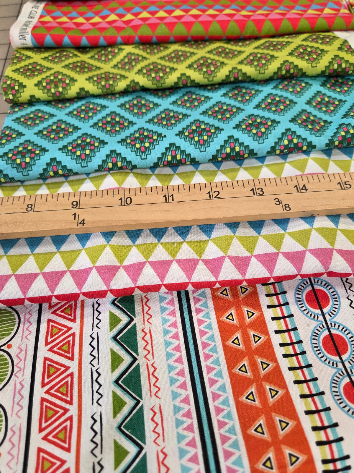 Several colorful patterned fabric bolts hanging vertically next to a wooden ruler for scale, showing geometric and tribal-style prints in pink, blue, green, orange, yellow, and white.