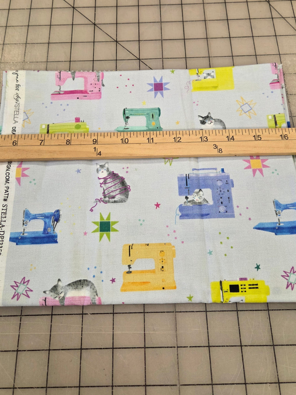 A piece of fabric with a white background and a colorful print of vintage sewing machines, stars, and cats, laid flat on a gridded cutting mat with a wooden ruler placed across it for scale.