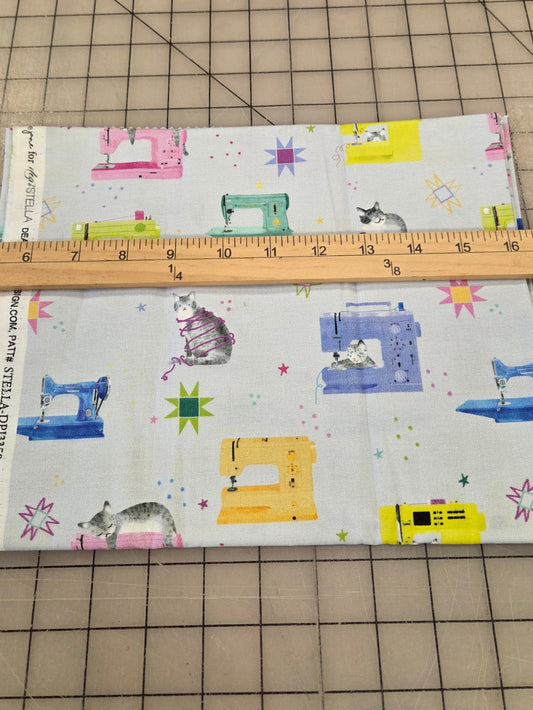 A piece of fabric with a white background and a colorful print of vintage sewing machines, stars, and cats, laid flat on a gridded cutting mat with a wooden ruler placed across it for scale.