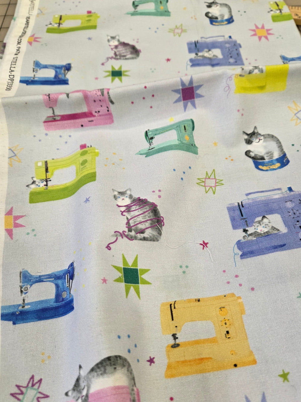 A folded piece of white fabric with a colorful print of sewing machines, cats, and starbursts, displayed on a cutting mat.