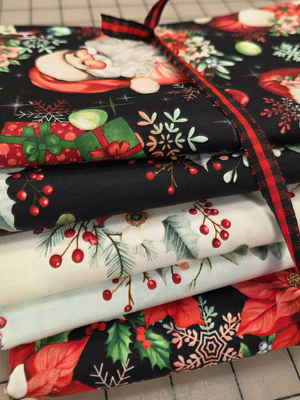 A stack of folded Christmas-themed fabric pieces tied with a red and black plaid ribbon, showing holiday prints including Santa, poinsettias, holly, berries, and snowflakes on a gridded cutting mat background.
