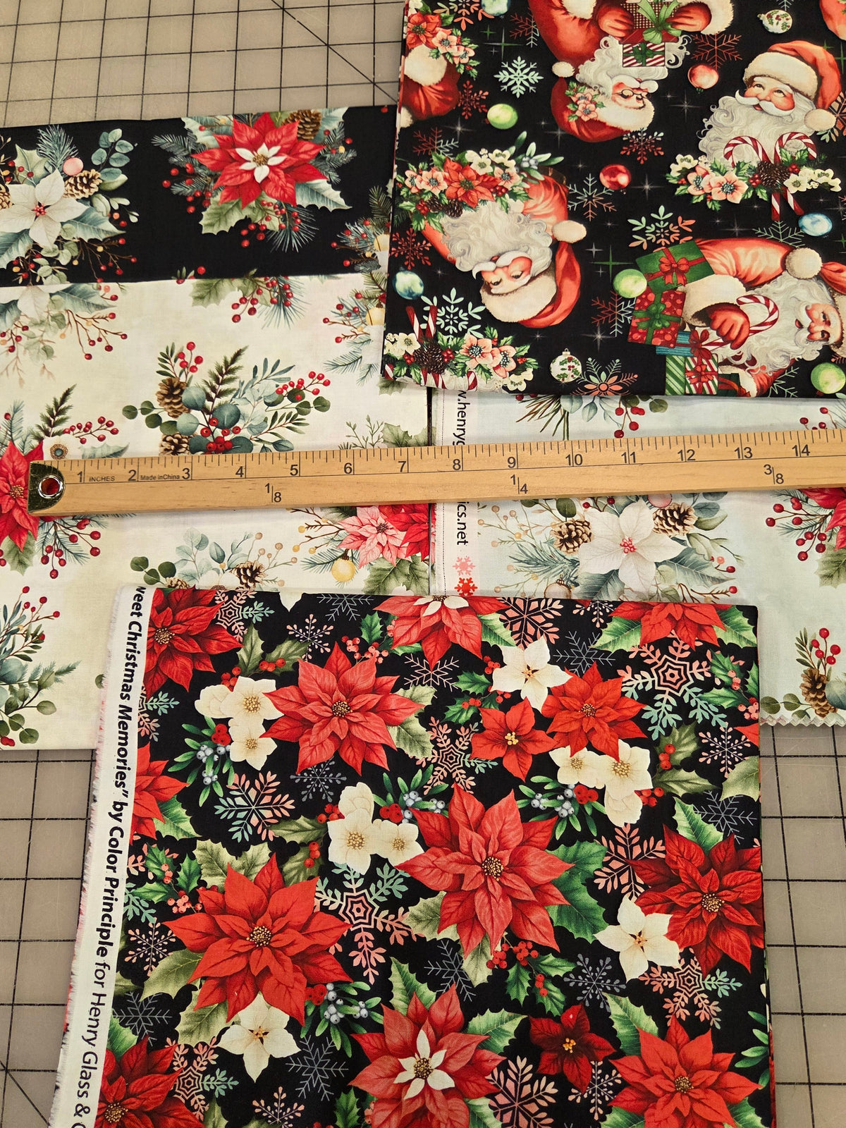 Three square fabric swatches with Christmas holiday prints are laid out on a gridded cutting mat with a wooden ruler in the center. The fabrics feature Santa Claus, poinsettias, and floral motifs in red, green, white, and black.