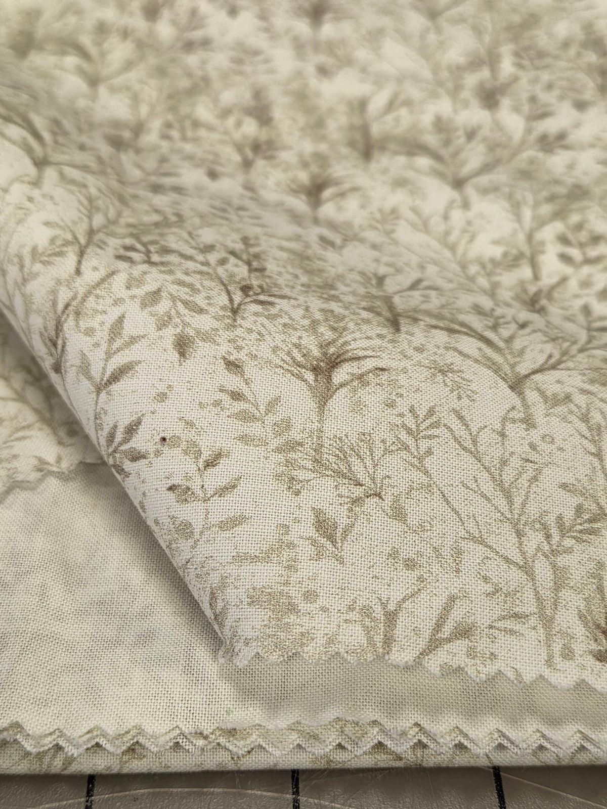 A close-up of a folded cream-colored fabric with a delicate beige floral and leaf pattern and a scalloped edge, placed next to a gridded cutting mat.