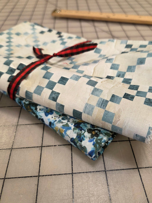 Two folded pieces of fabric on a gridded cutting mat. The top fabric is off-white with a blue-gray checkerboard pattern tied with a red and black ribbon, and the bottom fabric has a blue floral print.