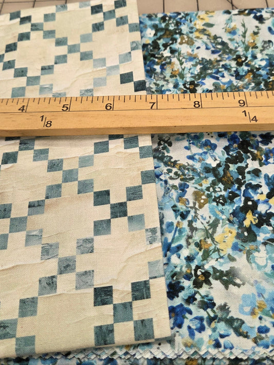Two folded pieces of patterned fabric laid on a gridded cutting mat with a wooden ruler across them for scale. One fabric has a blue and gray checkerboard pattern on an off-white base, and the other has a blue, gray, black, and yellow floral/abstract print on a white base.