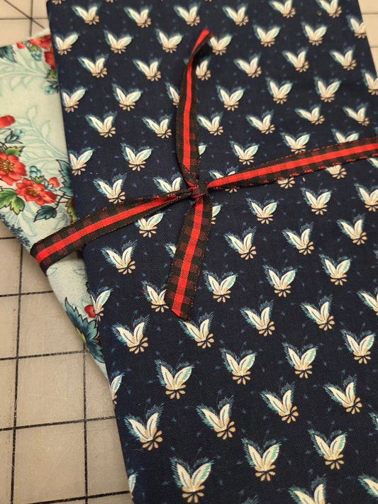 A folded dark navy fabric with a repeating light blue and beige butterfly pattern, tied with a red ribbon, resting on top of another floral-patterned fabric on a beige grid cutting mat.