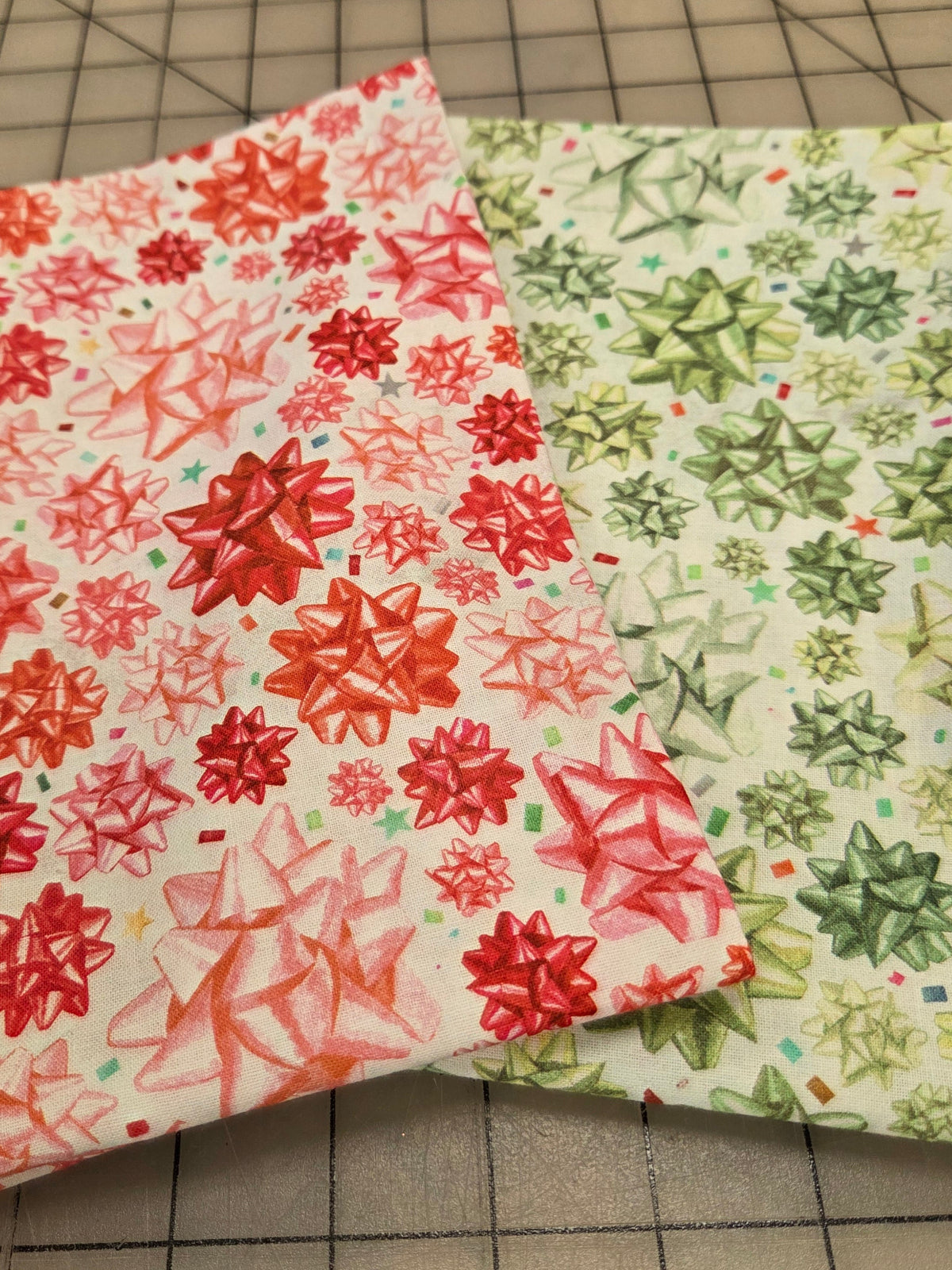 Two patterned fabric pieces with bow motifs, one in red and pink on white and one in green and yellow on white, displayed on a gridded cutting mat with a wooden ruler at the top.