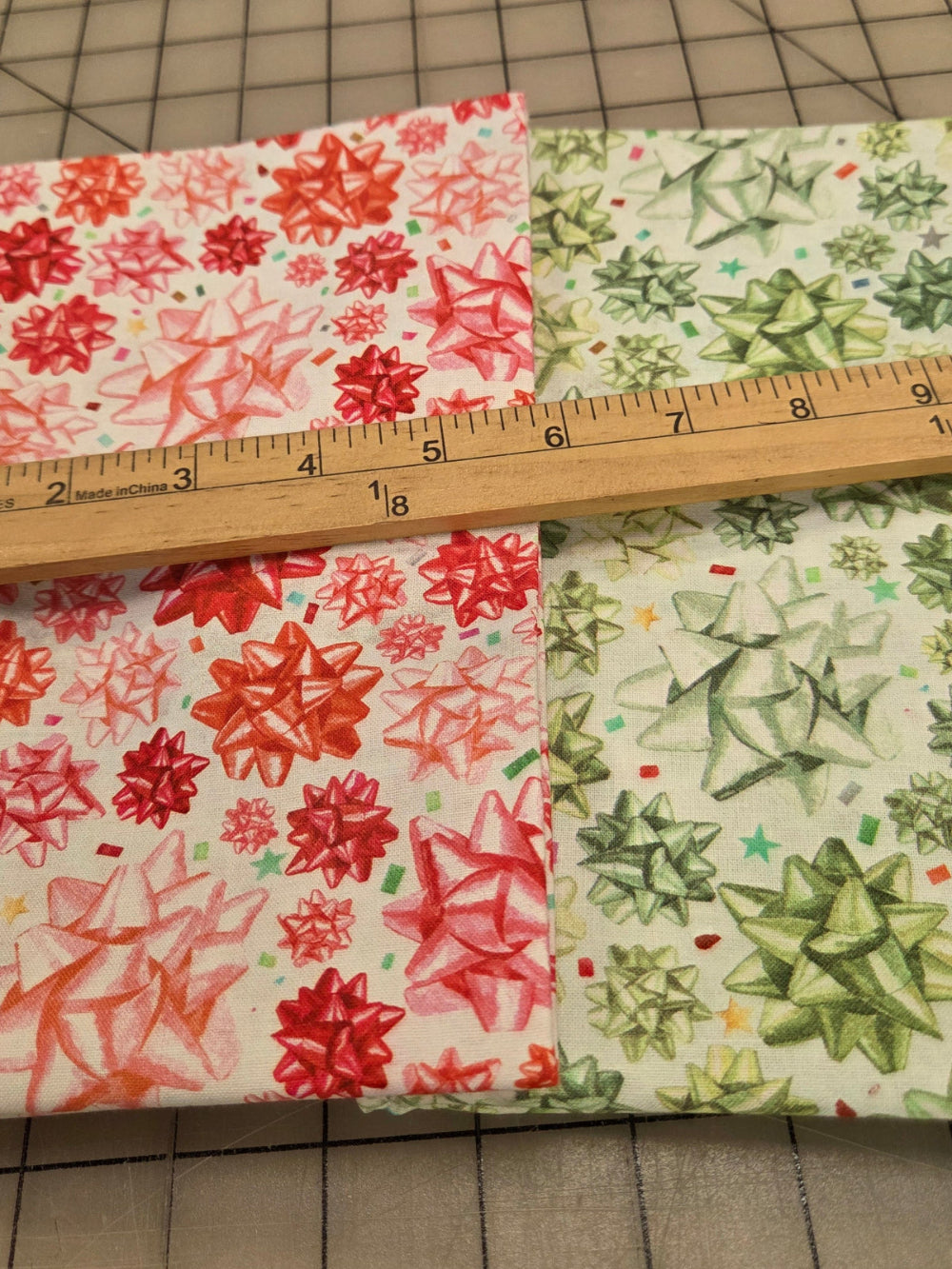Two folded pieces of patterned fabric with bow motifs, one in green and one in red, laid on a beige grid cutting mat with a wooden ruler for scale.