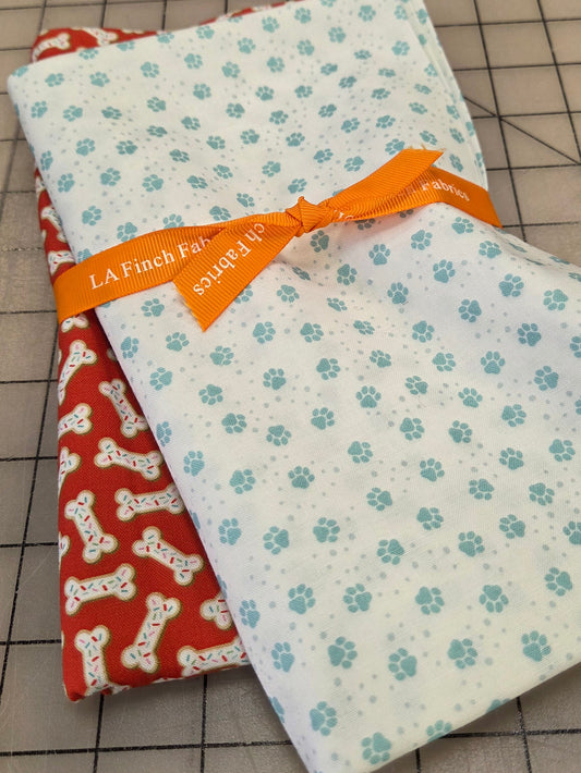 Two folded fabric pieces on a gridded cutting mat: a white fabric with small light blue paw prints tied with an orange ribbon, and a red fabric with white bone prints underneath.