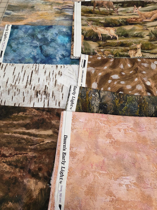 Six fabric swatches with printed labels are arranged on a gridded cutting mat. The fabrics feature various nature-themed prints including deer in a forest, birch trees, and textured earth tones, as well as a soft pink landscape.