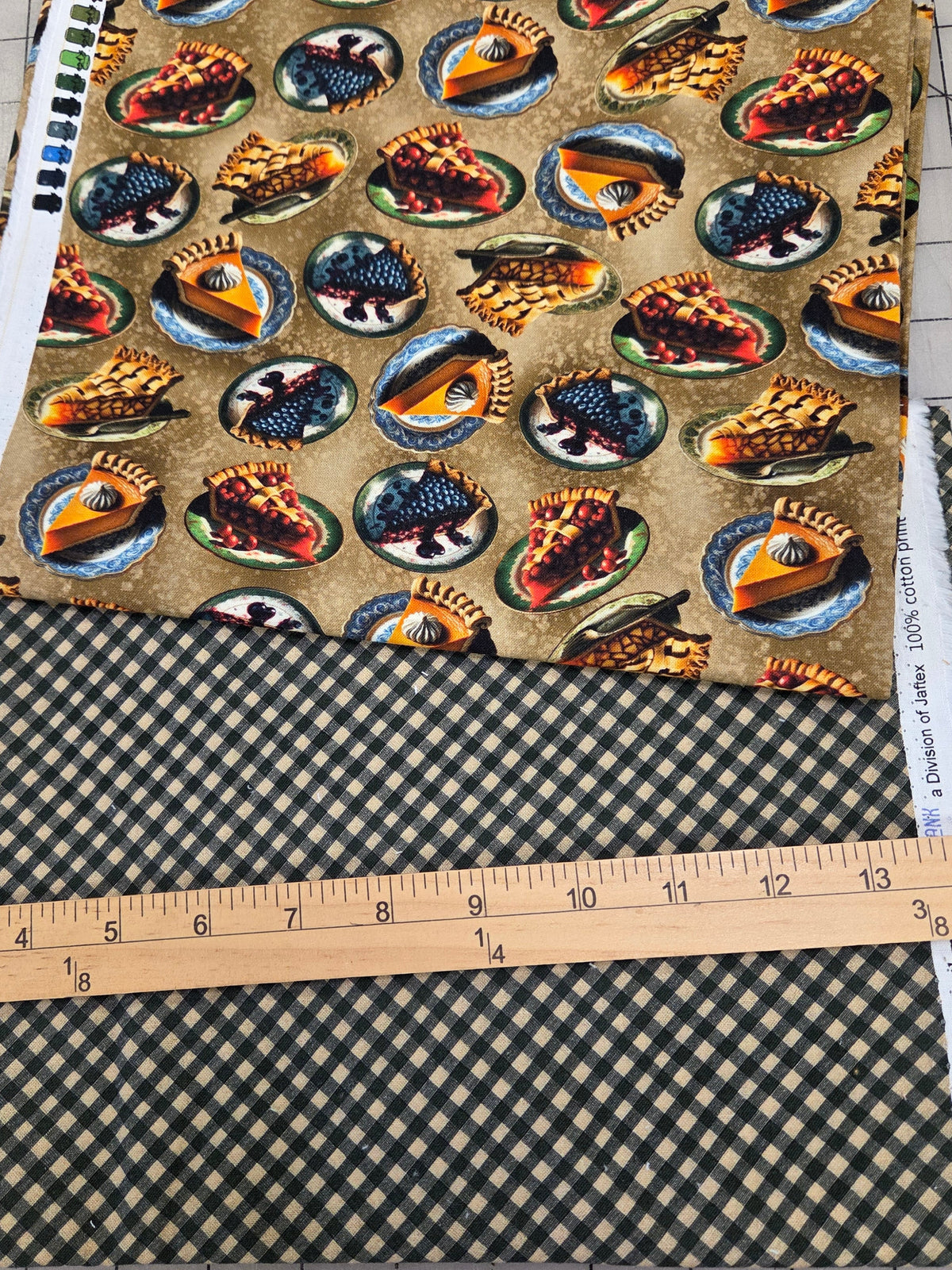 Two folded fabrics laid flat on a gridded cutting mat, with a wooden ruler in front for scale. The top fabric has a tan background with a repeating pattern of round medallions containing blue, red, yellow, and black motifs.