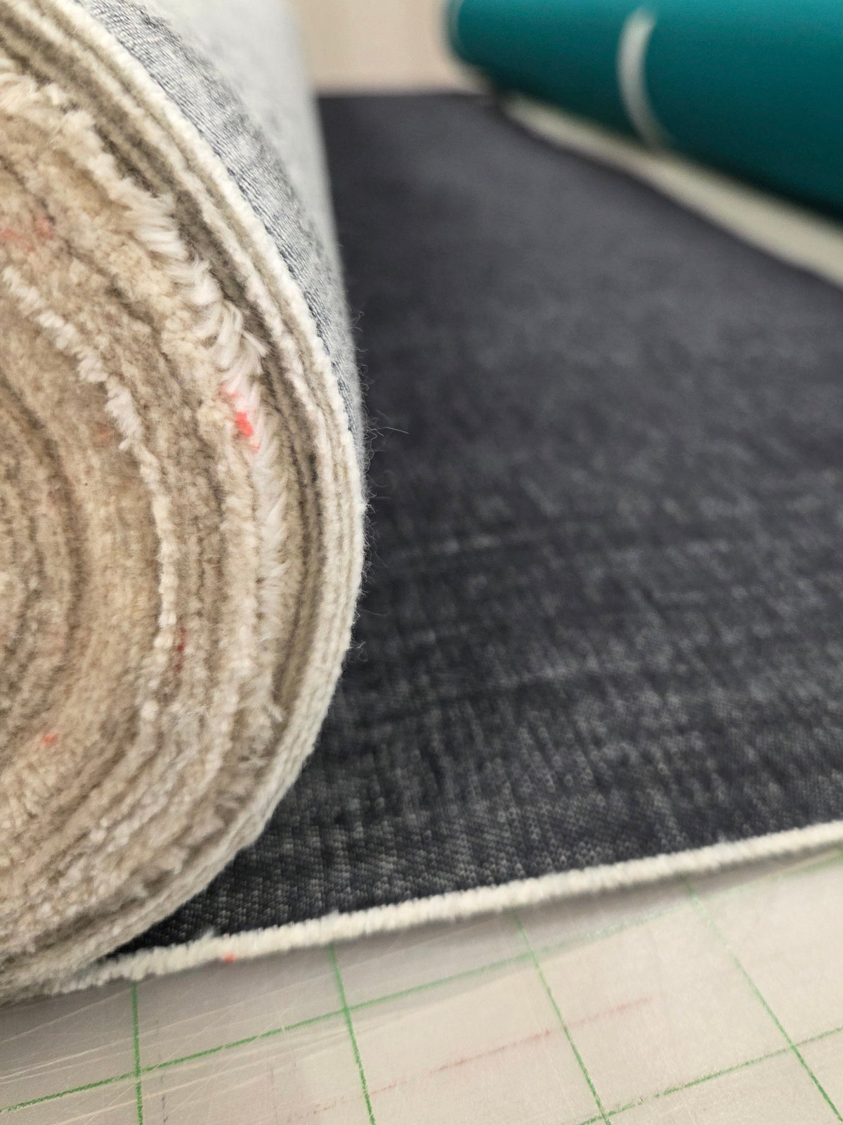 A close-up of a dark charcoal gray woven fabric roll unspooled onto a cutting mat, with other fabric rolls blurred in the background of what appears to be a sewing or fabric workshop.