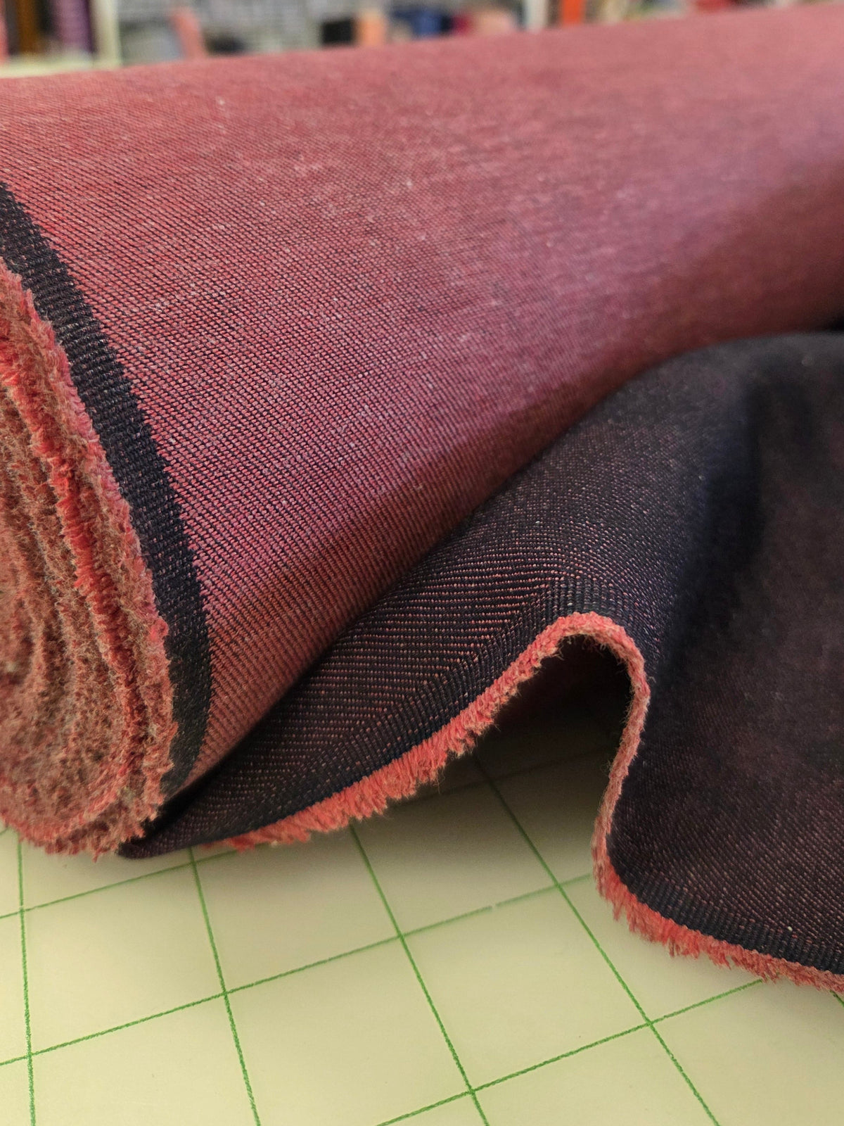 A close-up of a rolled piece of fabric with a dusty rose or mauve-pink front side and a dark charcoal gray reverse side, resting on a green-lined cutting mat with shelves of other fabrics blurred in the background.