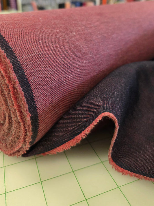 A close-up of a rolled piece of fabric with a dusty rose or mauve-pink front side and a dark charcoal gray reverse side, resting on a green-lined cutting mat with shelves of other fabrics blurred in the background.