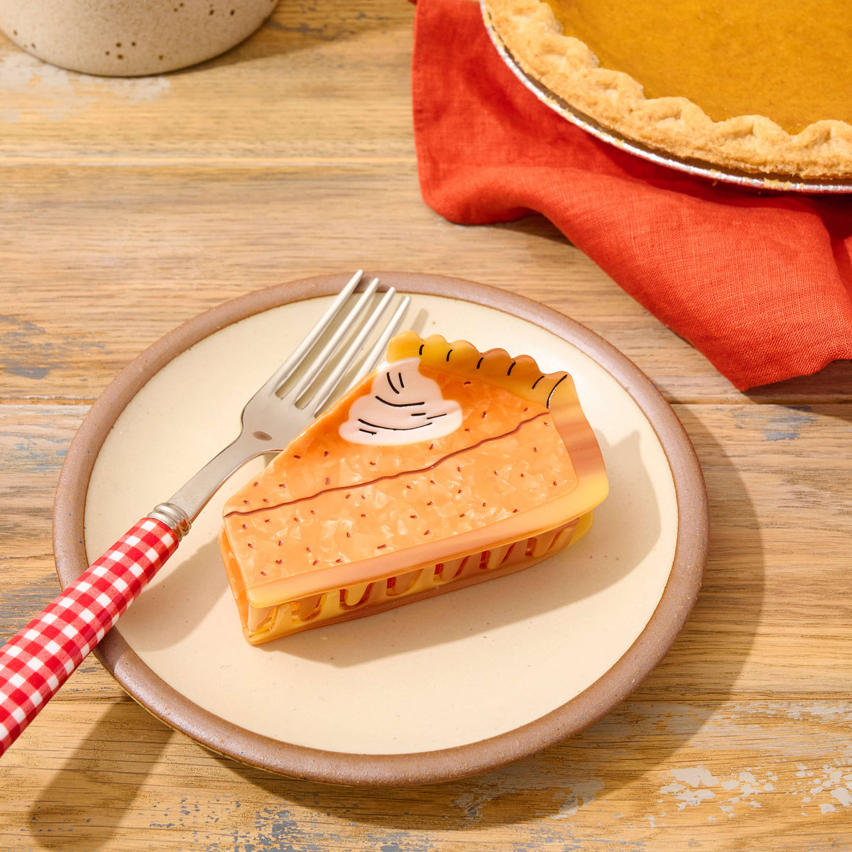 A ceramic plate with a brown rim holds a slice of pumpkin pie with a decorative crust and a small whipped cream dollop. A fork with a red-and-white gingham handle rests on the plate.