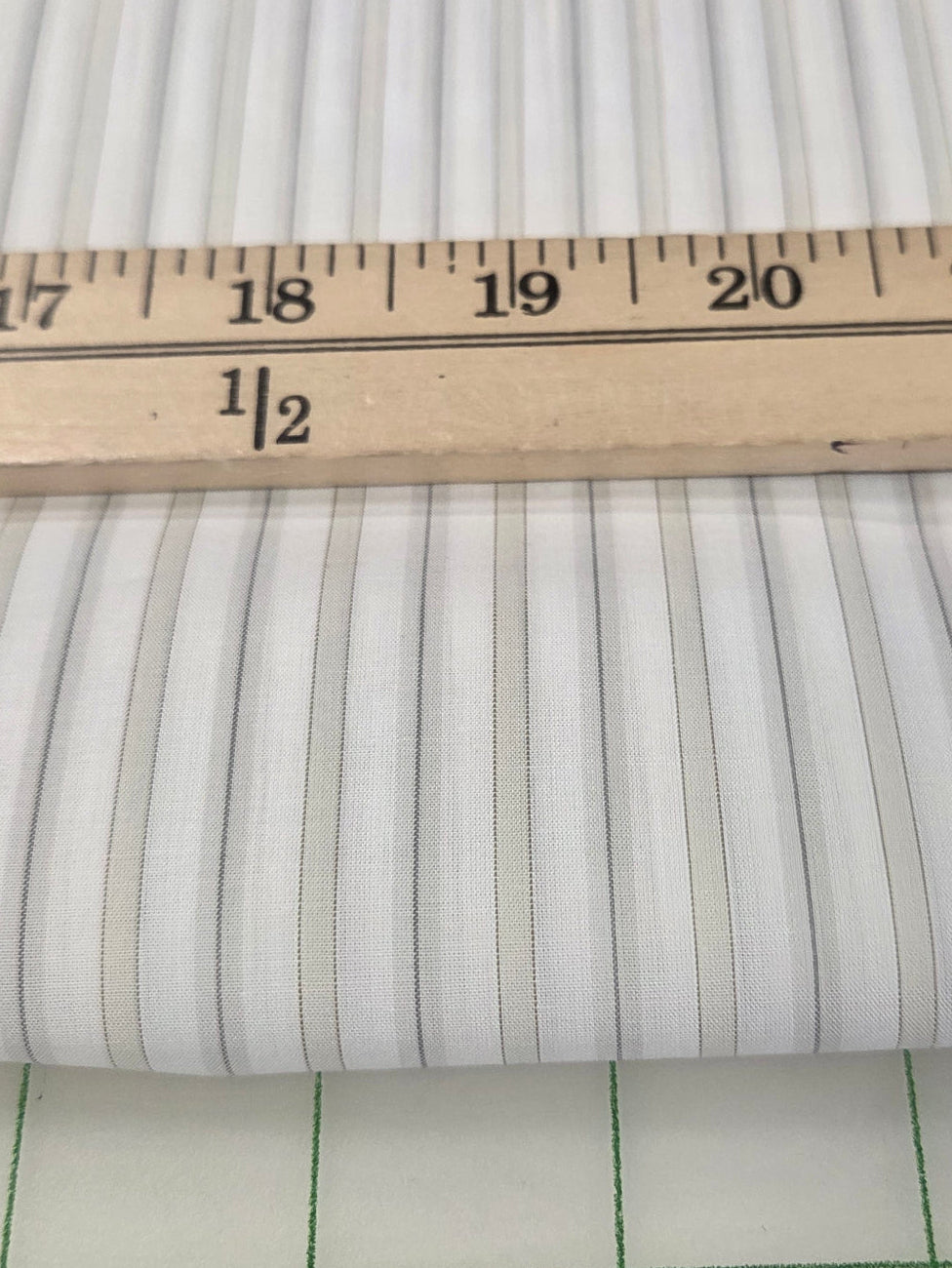A close-up of striped fabric with alternating white and gray vertical stripes, shown with a wooden ruler for scale on top and a gridded cutting mat below.