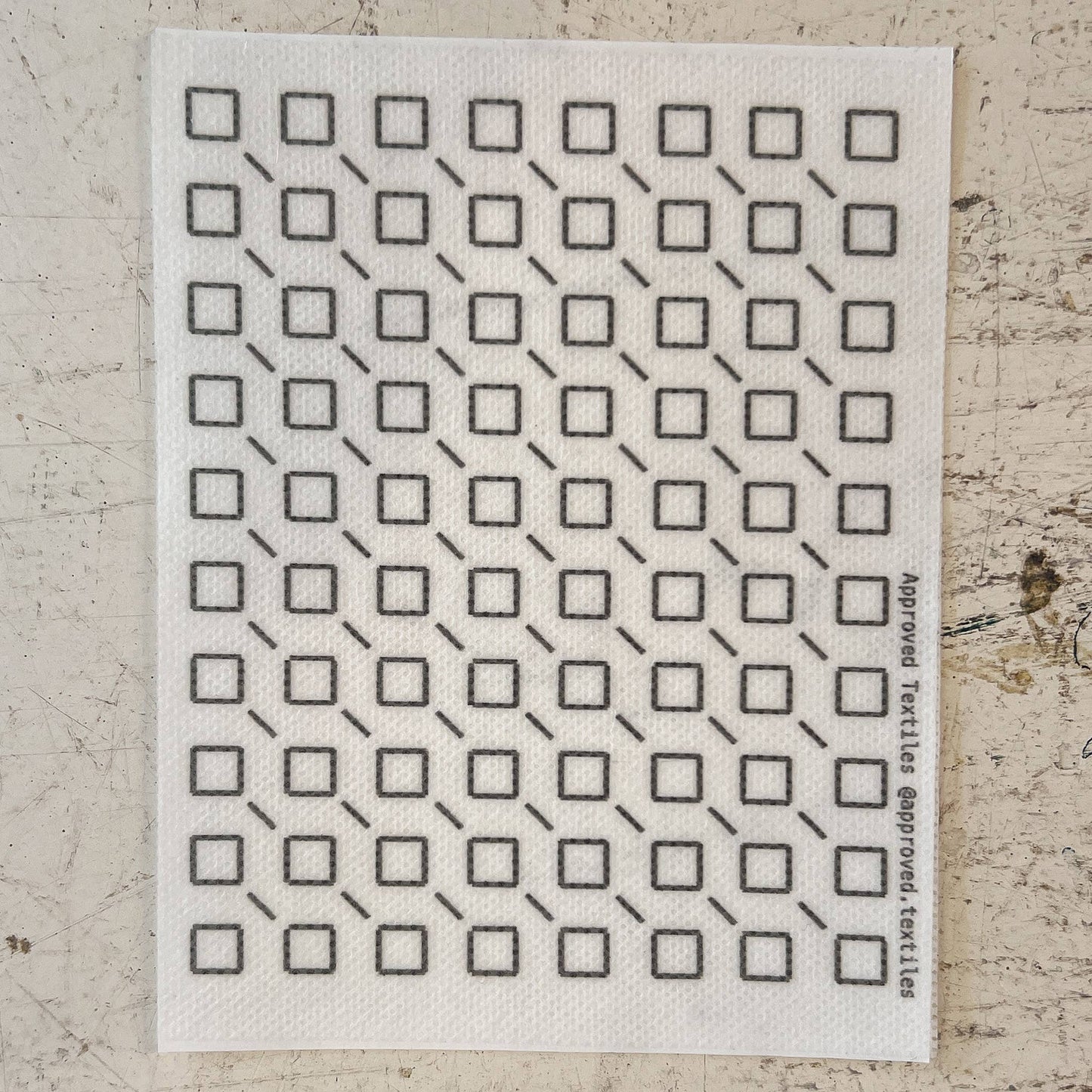 White cloth with black geometric pattern on a textured beige surface. mending supplies