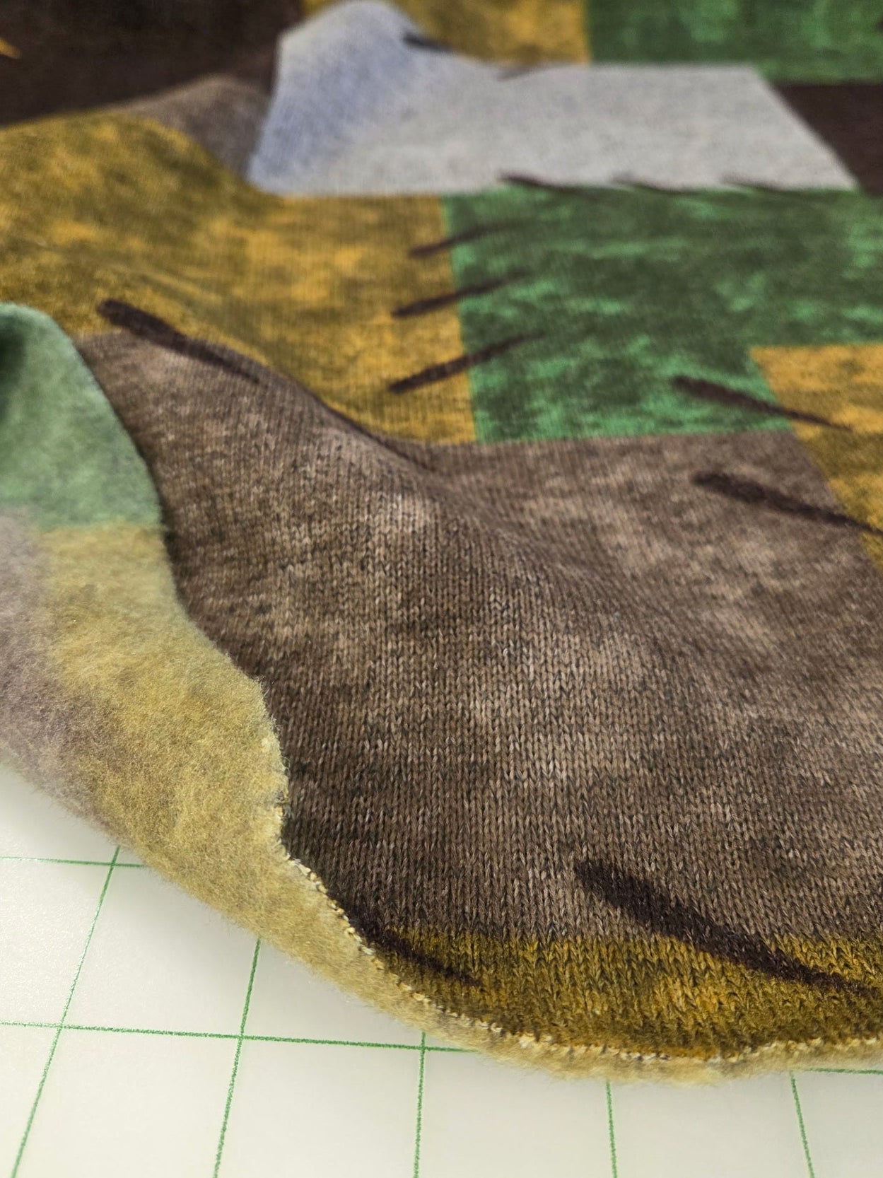 Close-up of a patchwork fabric with green, brown, and yellow colors on a white grid background.