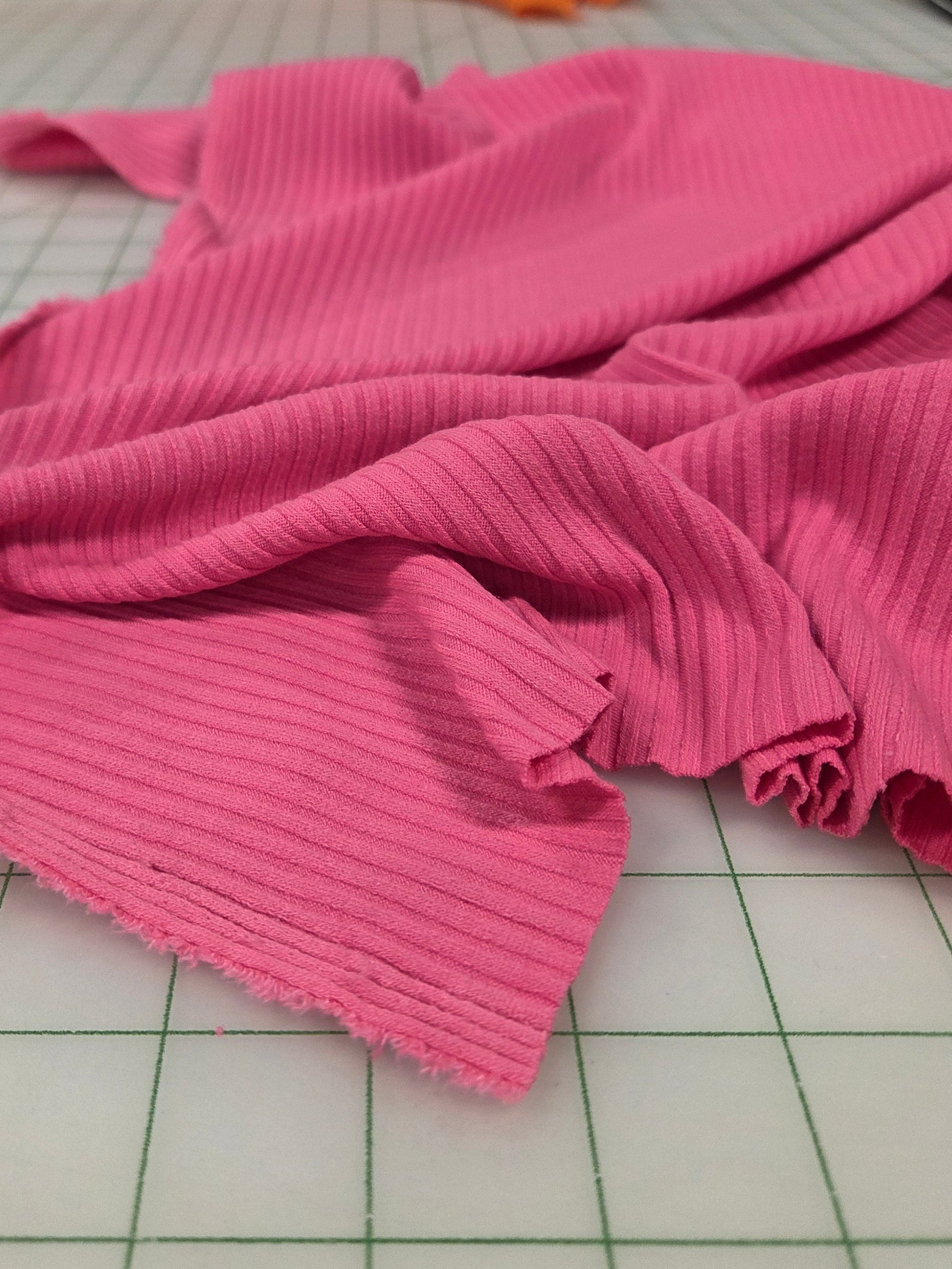 Pink fabric on a cutting mat with colorful fabrics in the background. Deadstock fabric. rib knit. poorboy rib. vibrant pink solid stretchy/