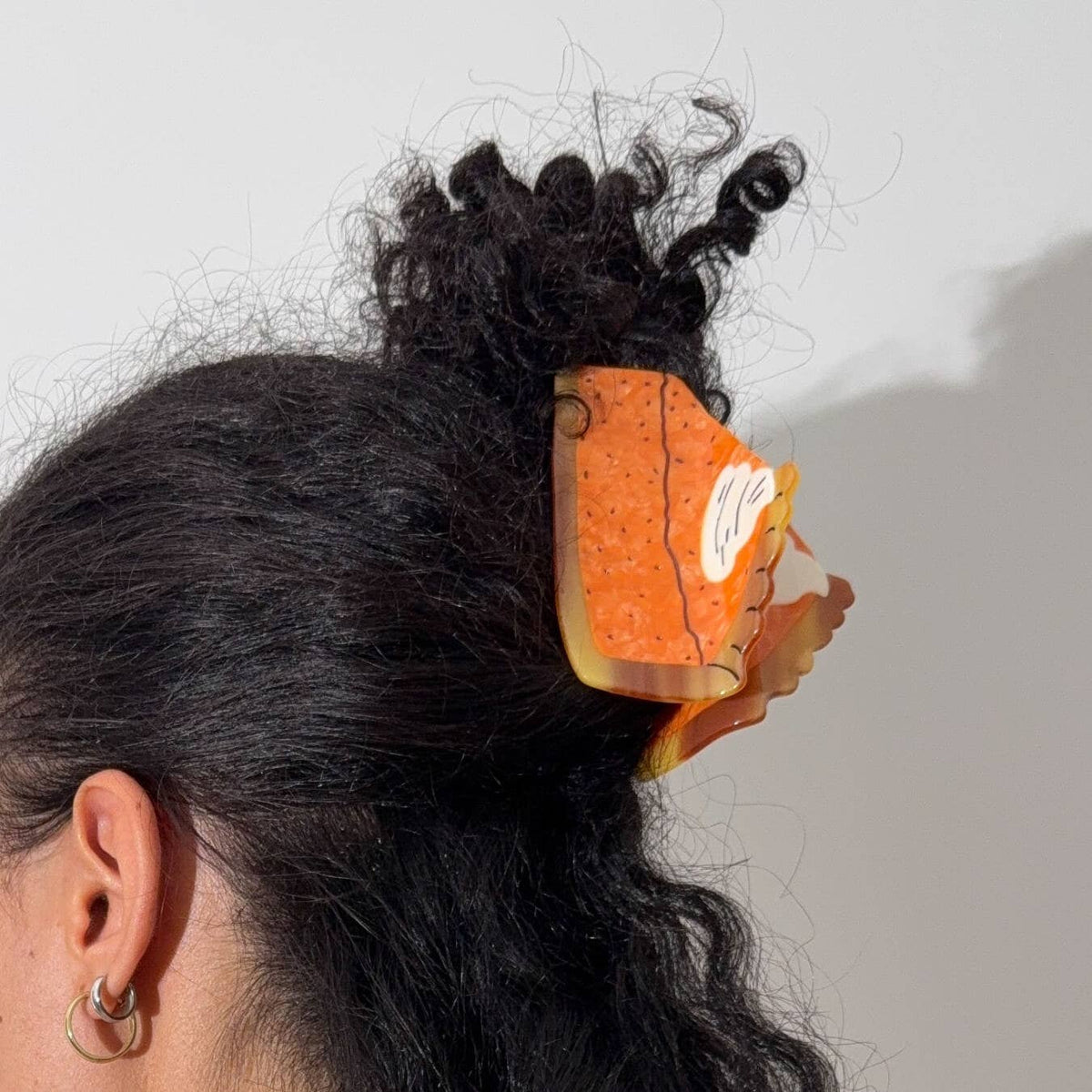 A close-up lifestyle shot of a person with curly black hair wearing a large, decorative orange hair claw clip with a white graphic illustration.